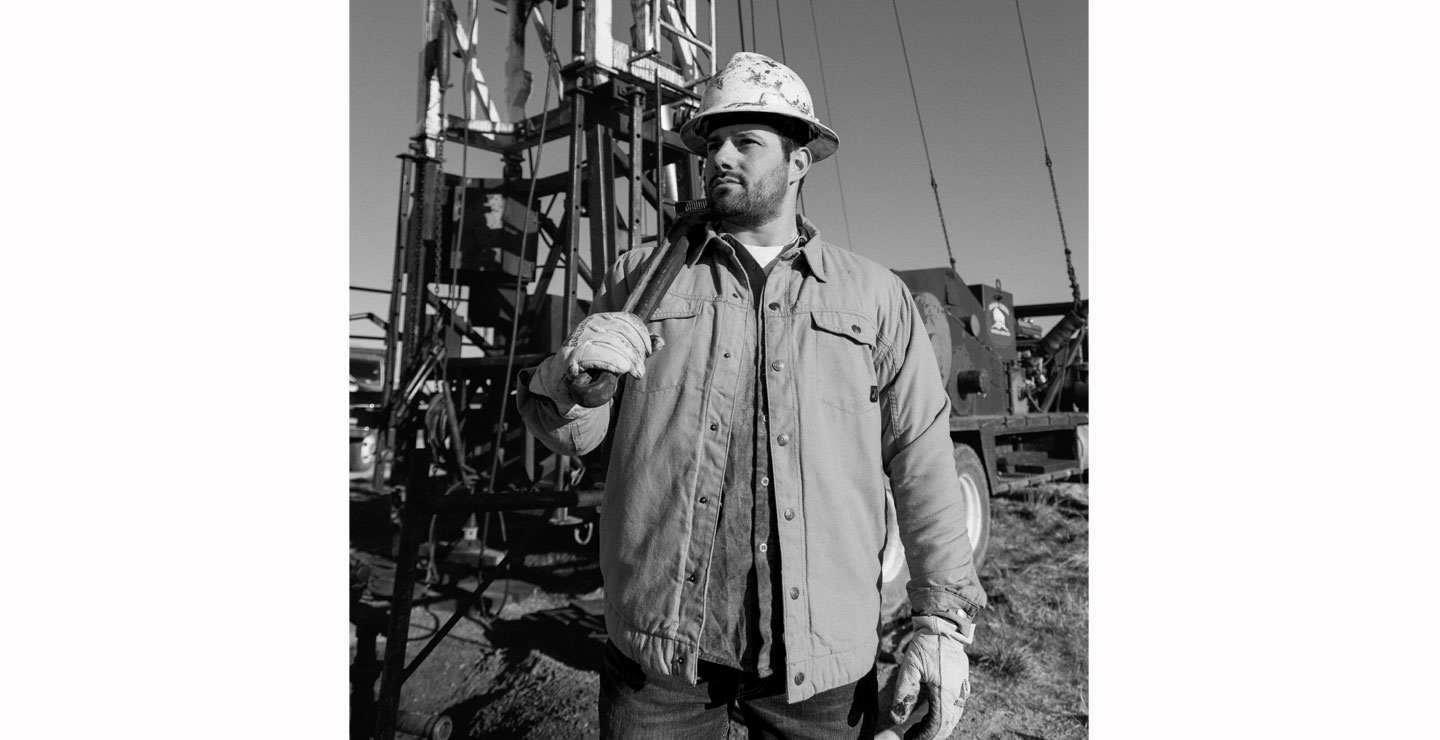 Una fotografía en blanco y negro de un hombre sosteniendo una llave inglesa, posando frente a una plataforma petrolera.